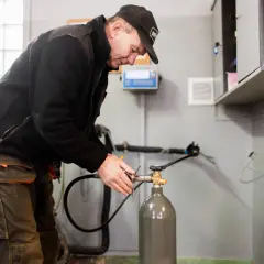 Pracownik w czapce i roboczych spodniach dokonuje napełniania butli CO2, podłączając wąż do zaworu.