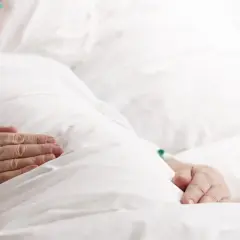 Dłonie starszej osoby spoczywają na białej pościeli, czuć delikatny oddech biota.