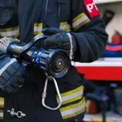 Strażak w rękawicach trzyma prądownicę, gotowy do akcji. W tle wóz strażacki z wyposażeniem.