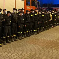 Strażacy w czarnych mundurach stoją w szeregu przed wozami strażackimi. Wśród nich można rozpoznać komendanta głównego PSP.
